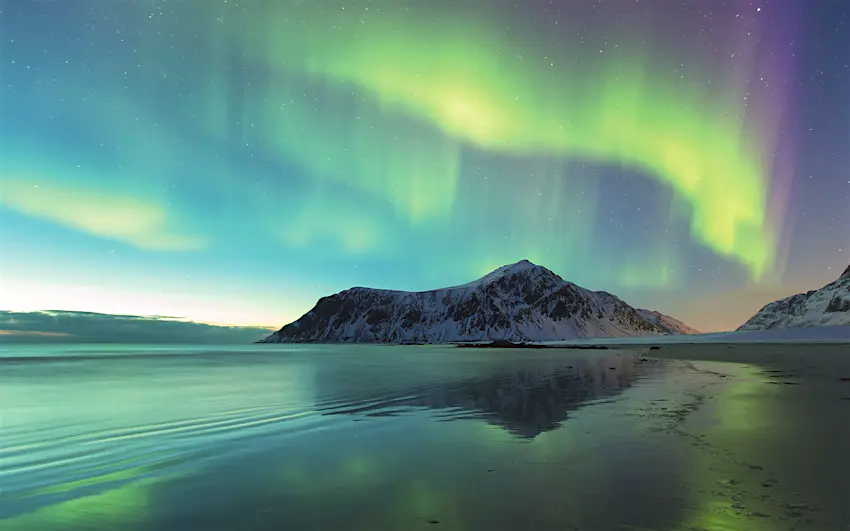 Features - "Northern Lights on Skagsanden Norway" Green waves of the aurora move over a beach in Norway. A snowy mountain is in the background. © Spreephoto.de / Getty Images