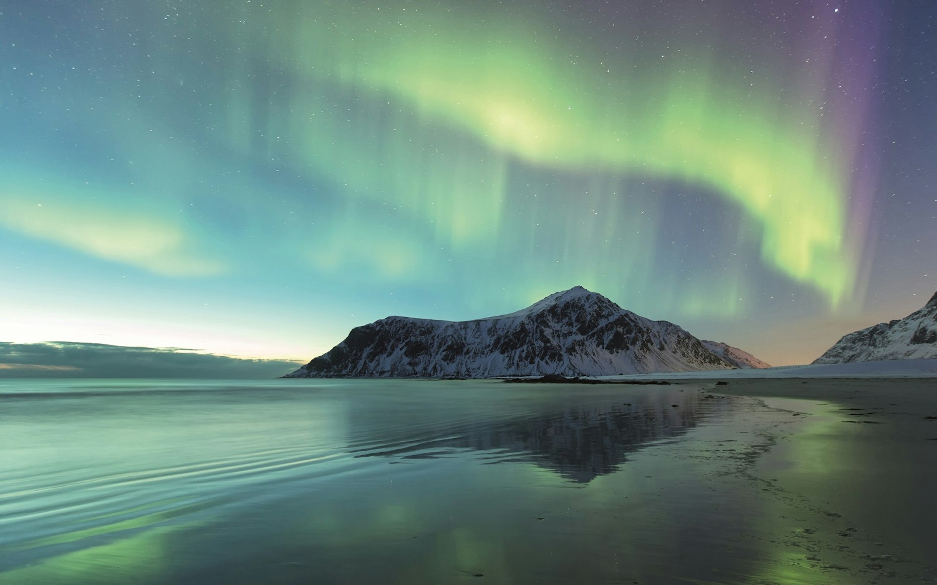 Green waves of the aurora move over a beach in Norway. A snowy mountain is in the background. © Spreephoto.de / Getty Images