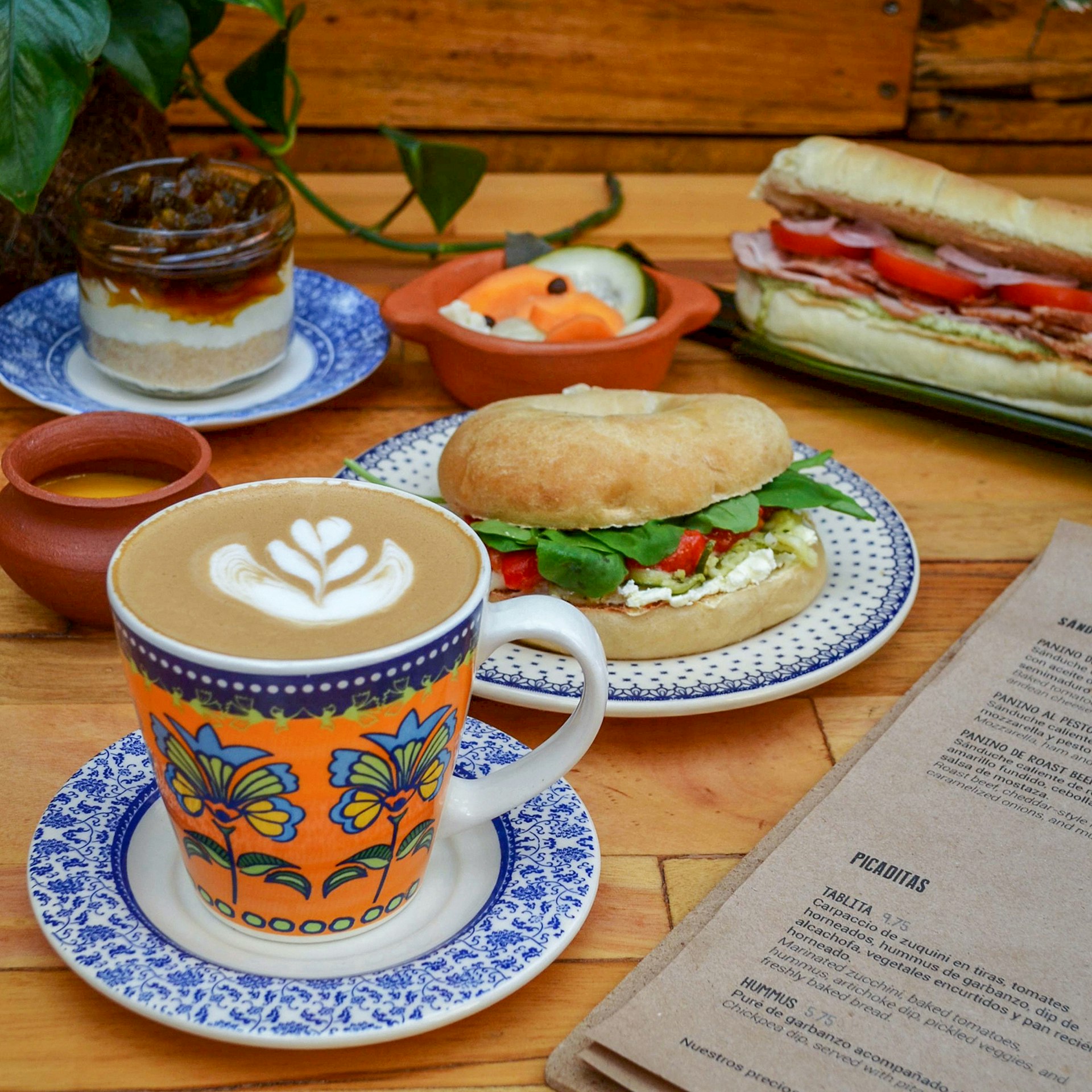 A wooden table set with a coffee, two sandwiches and a dessert, with the corner of a menu © Botánica