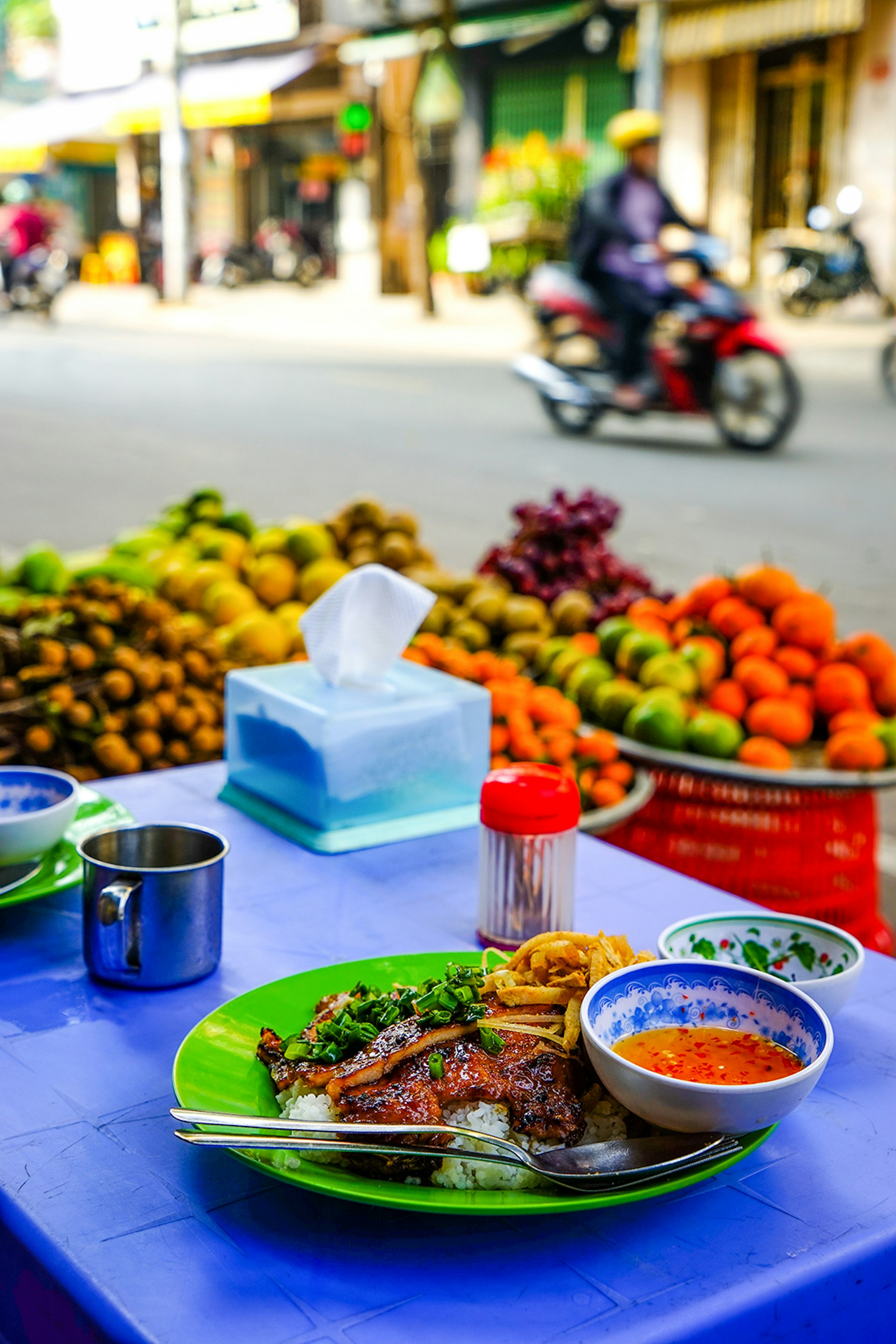 A bowl of food on a blue tablecloth in the foreground with fruit and a man on a motorbike in the background © James Pham / Lonely Planet