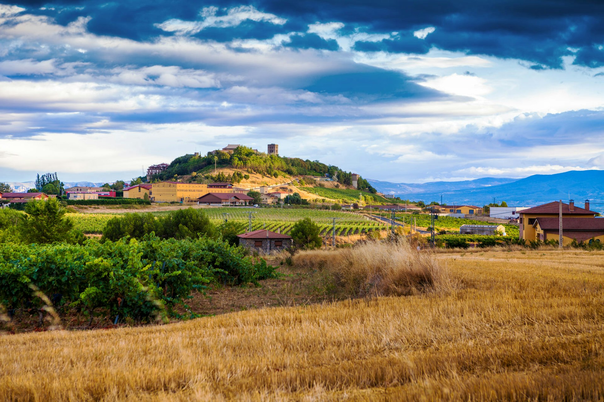 The vineyards of Rioja, Spain, with a town on a hill in the distance and a moody grey and blue sky. Where to go in October