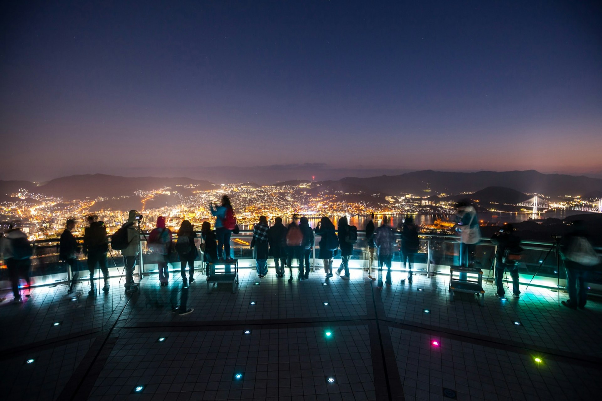 Tourists are silhouetted at night at an overlook, with the lights of Nagasaki below © Putt Sakdhnagool / Getty Images 
