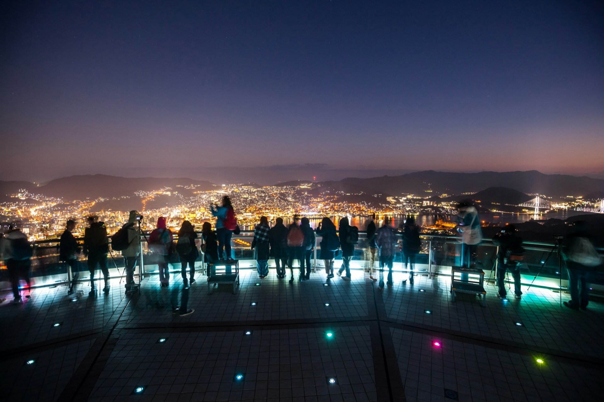Tourists are silhouetted at night at an overlook, with the lights of Nagasaki below © Putt Sakdhnagool / Getty Images