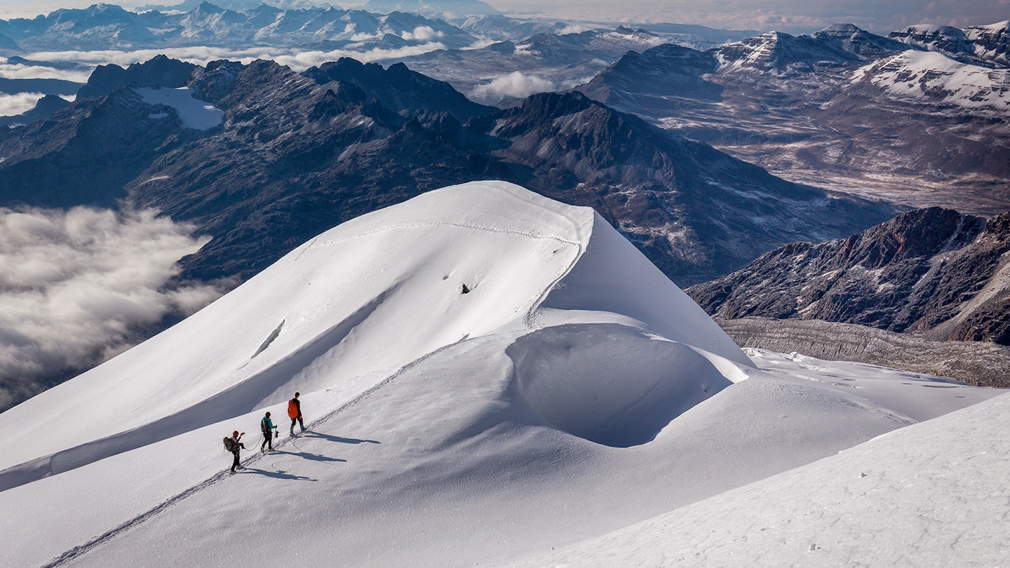 Three trekkers walk across a snowy slope with a rocky mountain range in the background © Subbotsky / Getty Images