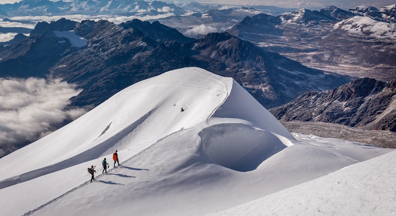 Three trekkers walk across a snowy slope with a rocky mountain range in the background © Subbotsky / Getty Images