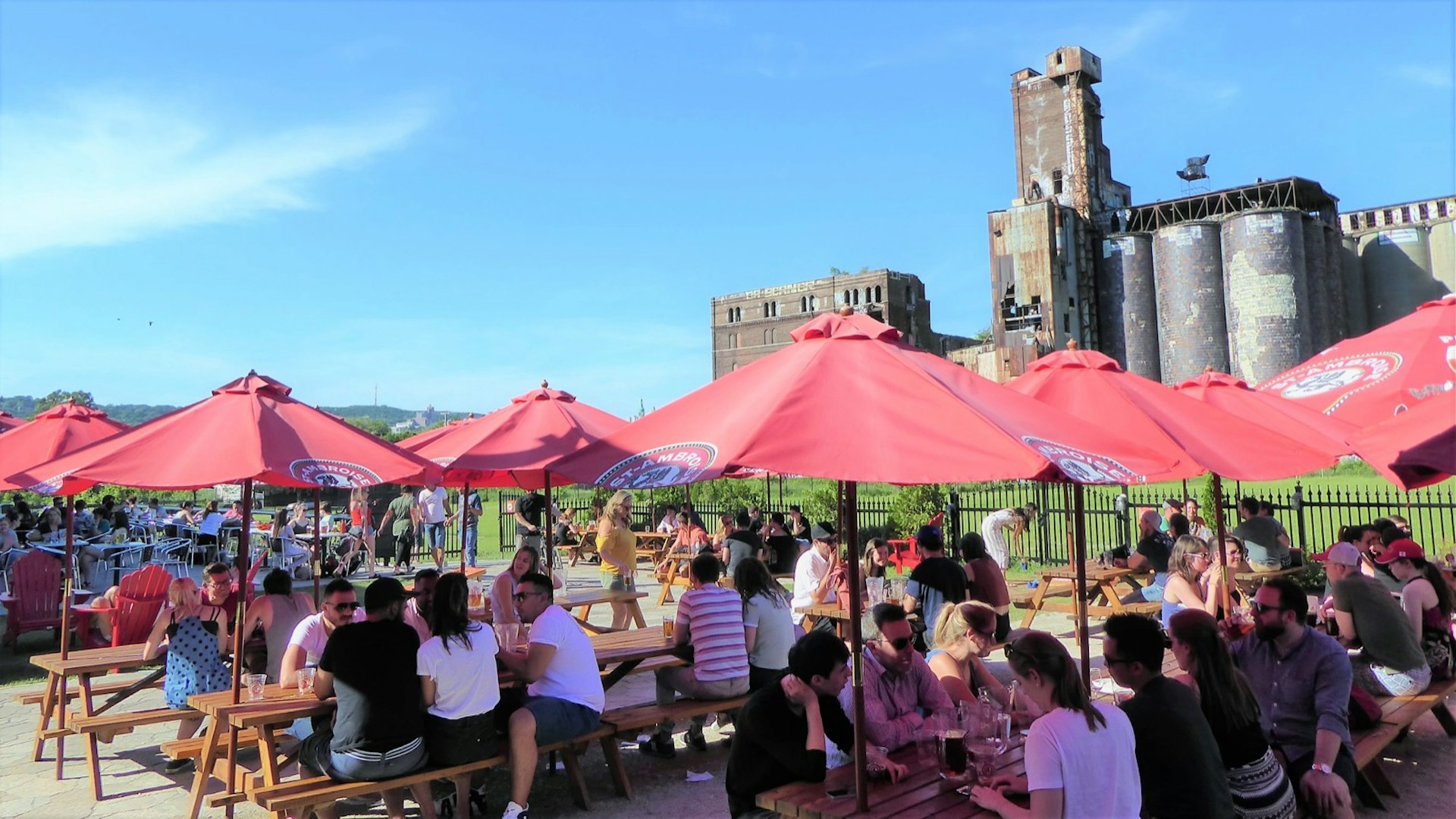 Several bright red umbrellas dominate a sunny patio with patrons drinking and socializing. In the distance is a dilapidated building © Jason Najum / Lonely Planet