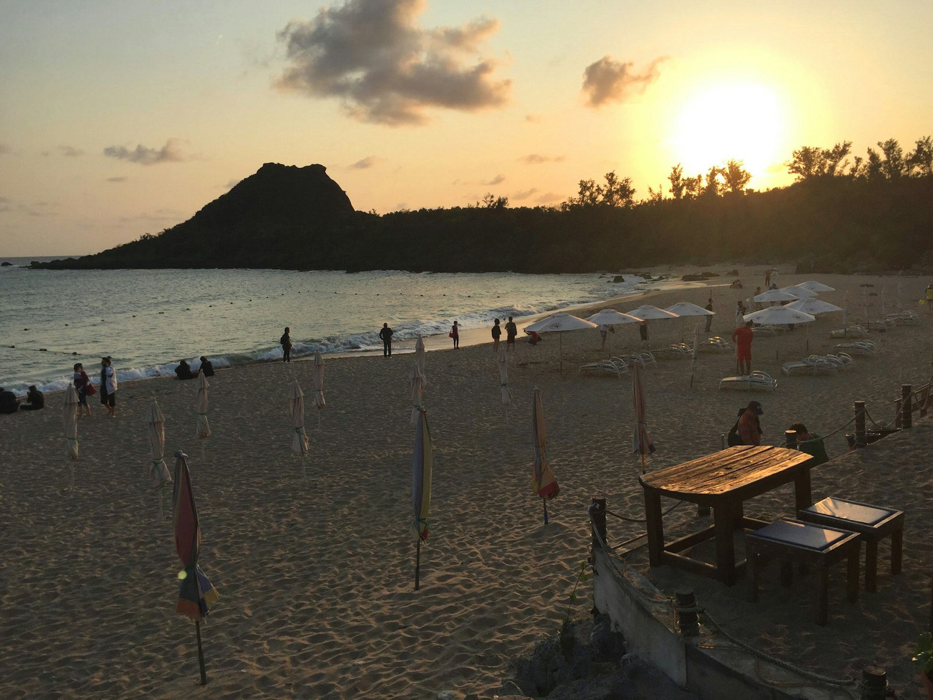 The sun sets over a small beach with a few people and some closed umbrellas. Superb swimming (and cocktails!), even in February, at Kenting's Little Bay Beach © Megan Eaves / Lonely Planet