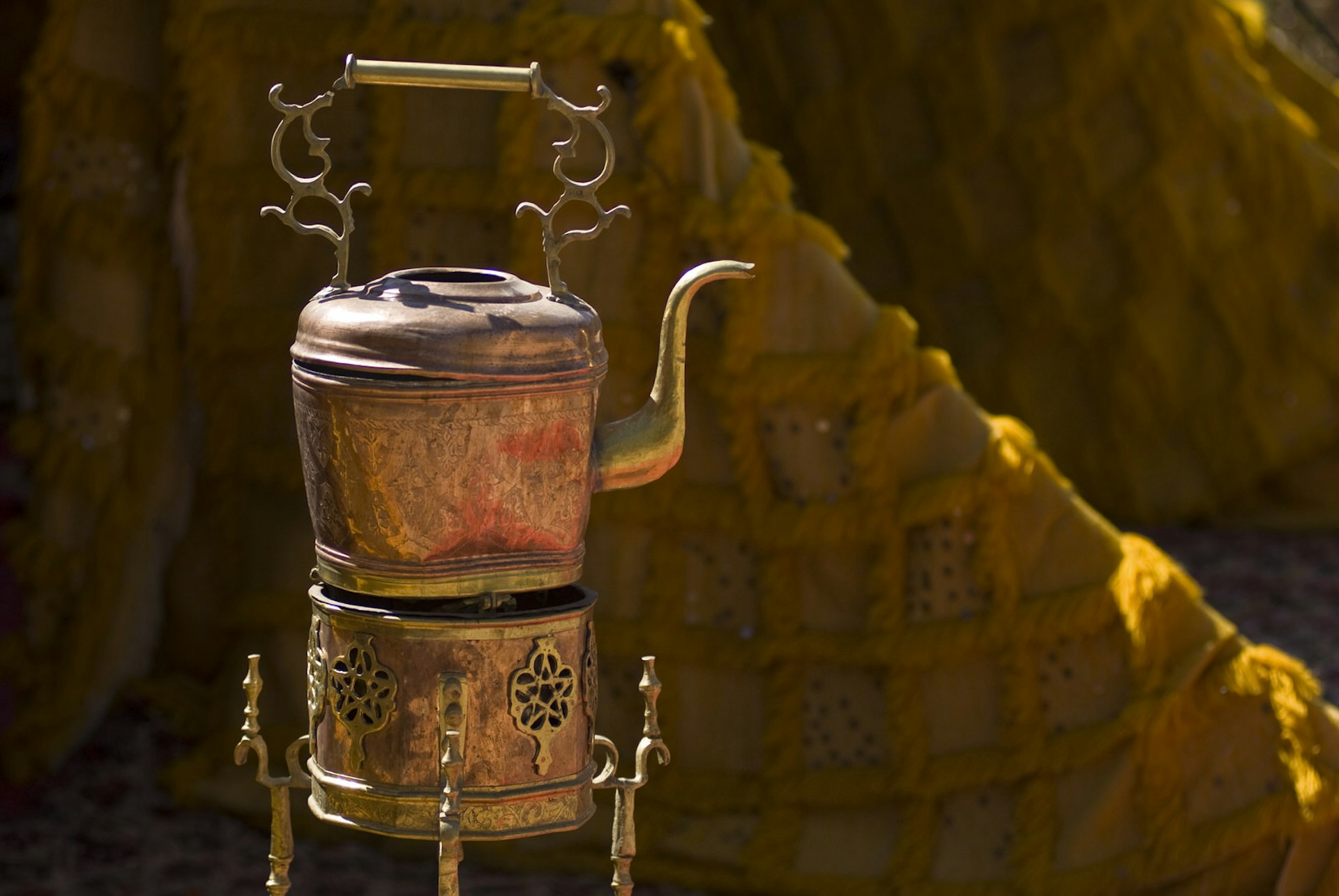 A highly decorative (and aged) copper teapot standing on an equally elaborate cooker.