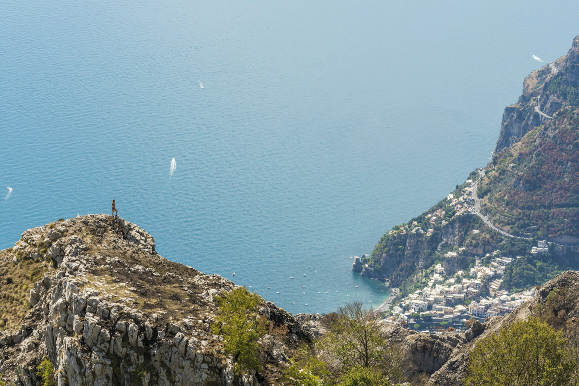 A hiker stands on a rocky promontory, looking down on a distant pastel-housed village with the sea below