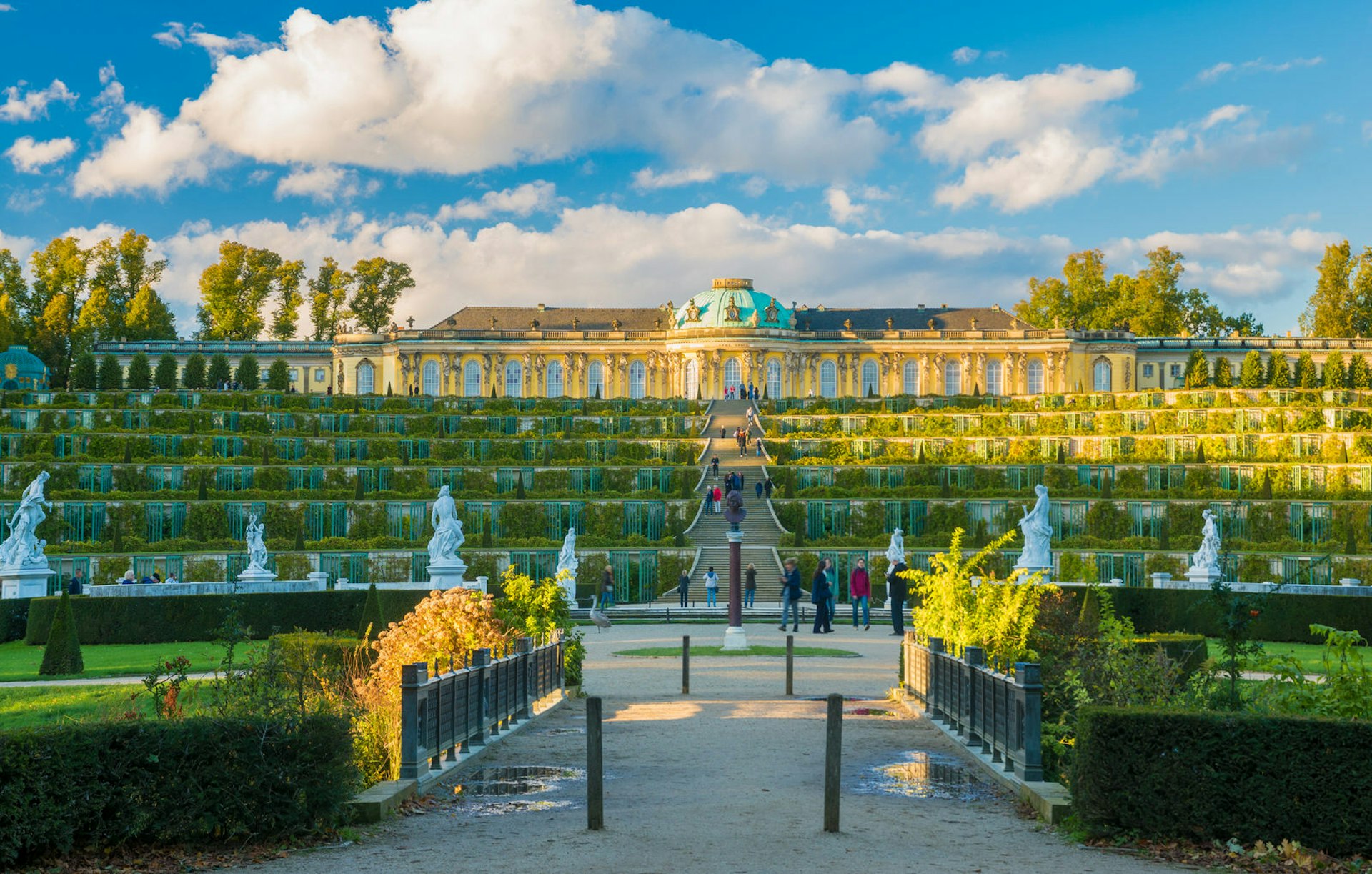 Berlin day trips - Sanssoucci Palace in Potsdam, which can be visited as a day trip from Berlin. The palace sits above a long flight of stairs which are flanked by statues and greenery. There is a copper-domed roof above the yellow walls and many curved windows. 