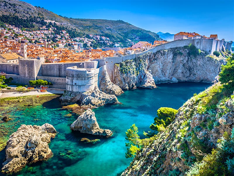 A view of the orange roofs of Dubrovnik, surrounded by stone city walls; the city is perched dramatically on cliffs above the bright turquoise Adriatic.
