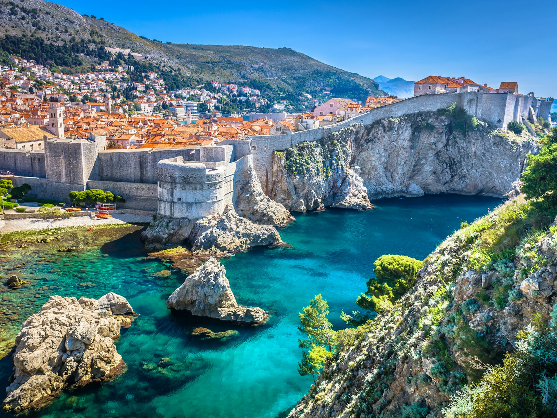 A high-angle view of Dubrovnik, a mass of roofs behind city walls perched above a stunning blue Adriatic; there is a mountainside beyond.