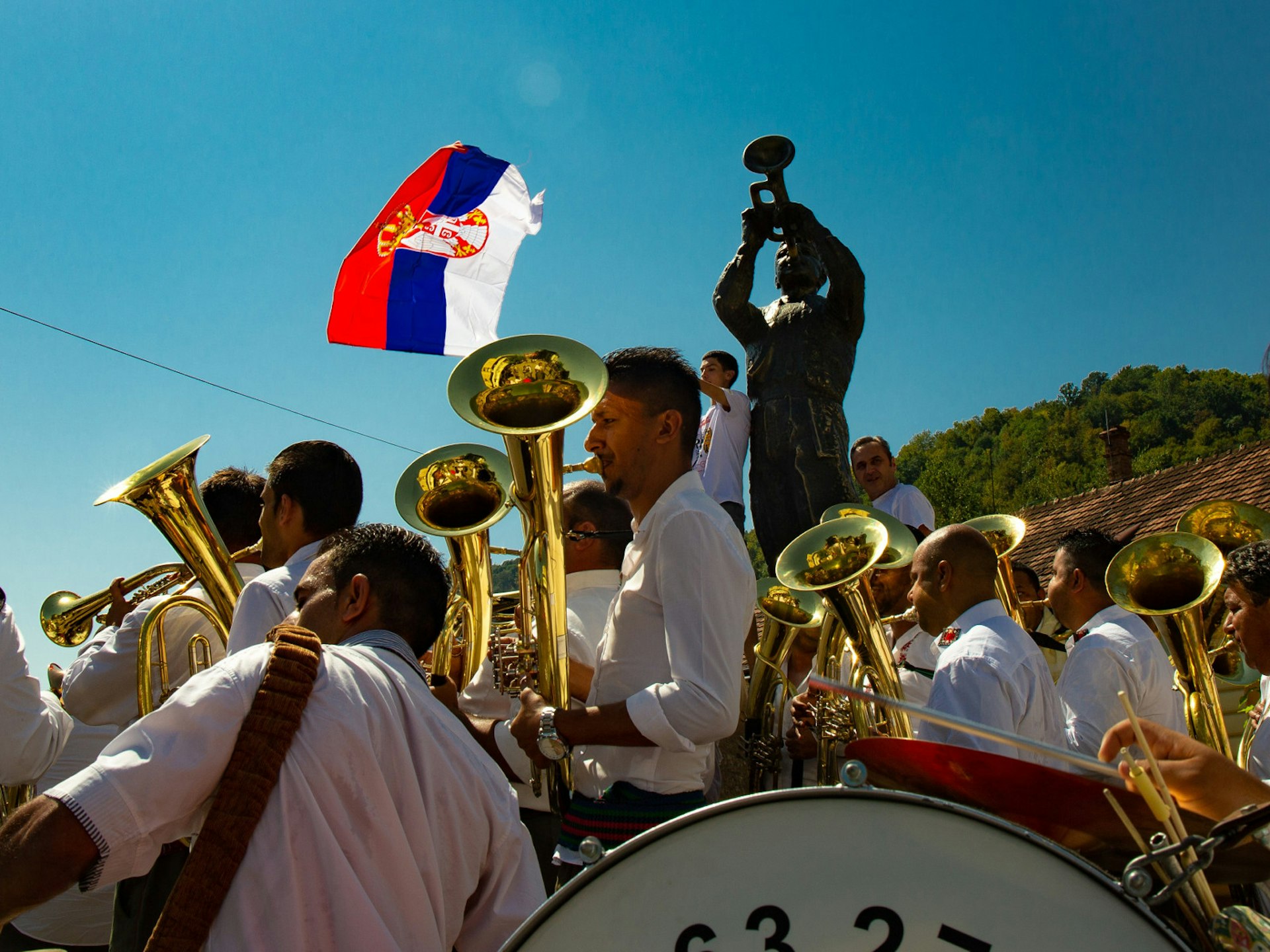 Roma brass bands parade through Guča during the 2017 Trumpet Festival in front of a blue sky as well as a statue of a trumpet player. Children stand at the base of the statue waving the Serbian flag © Aleksandar Donev / Lonely Planet