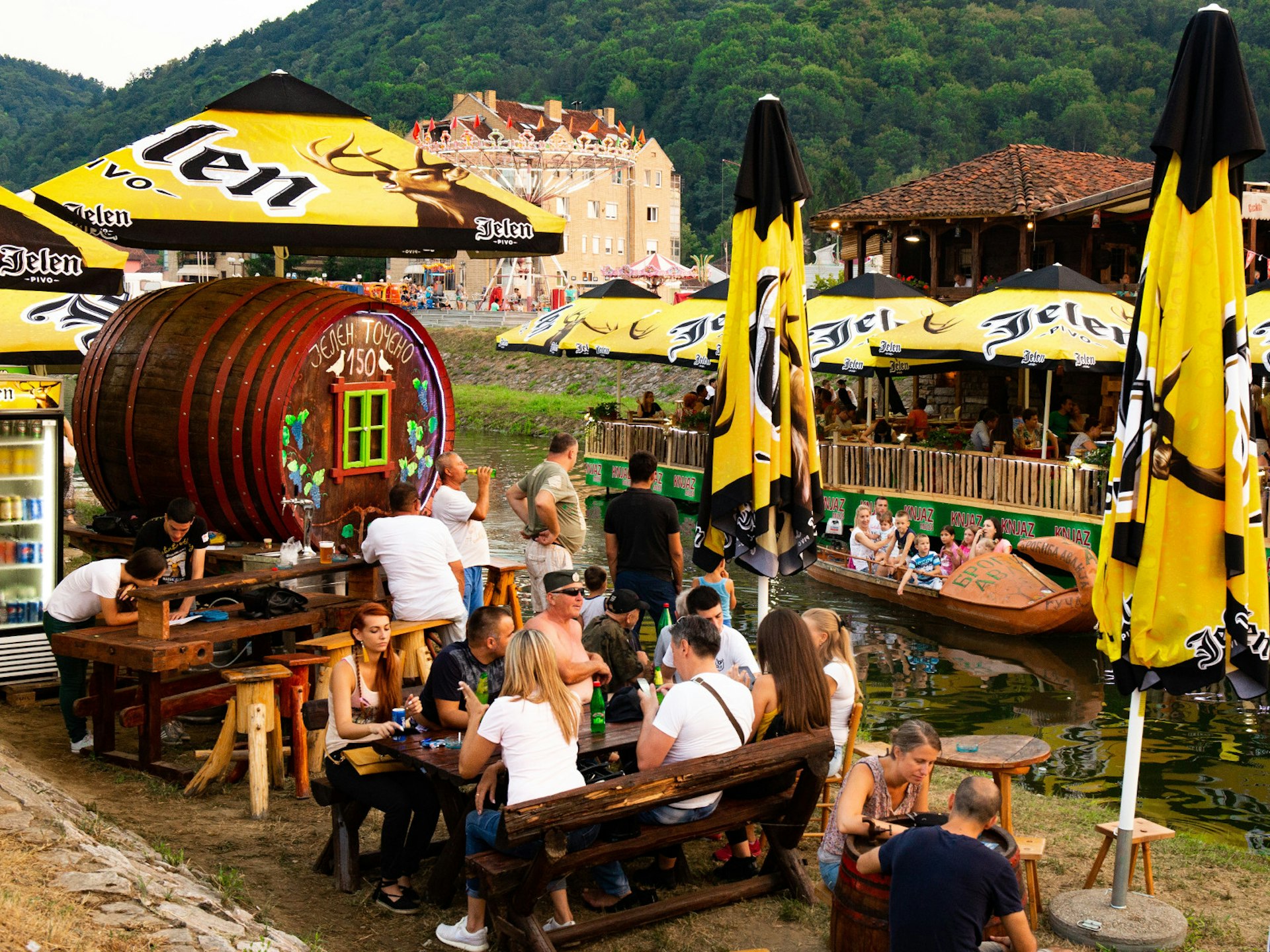 Outdoor riverside cafes packed with festival-goers in the village of Guča. Groups sit at picnic tables, under bright yellow umbrellas with rolling hills and forests in the background. A river runs between two cafes and festival goers are floating down it in a boat made to look like a huge clog © Aleksandar Donev / Lonely Planet