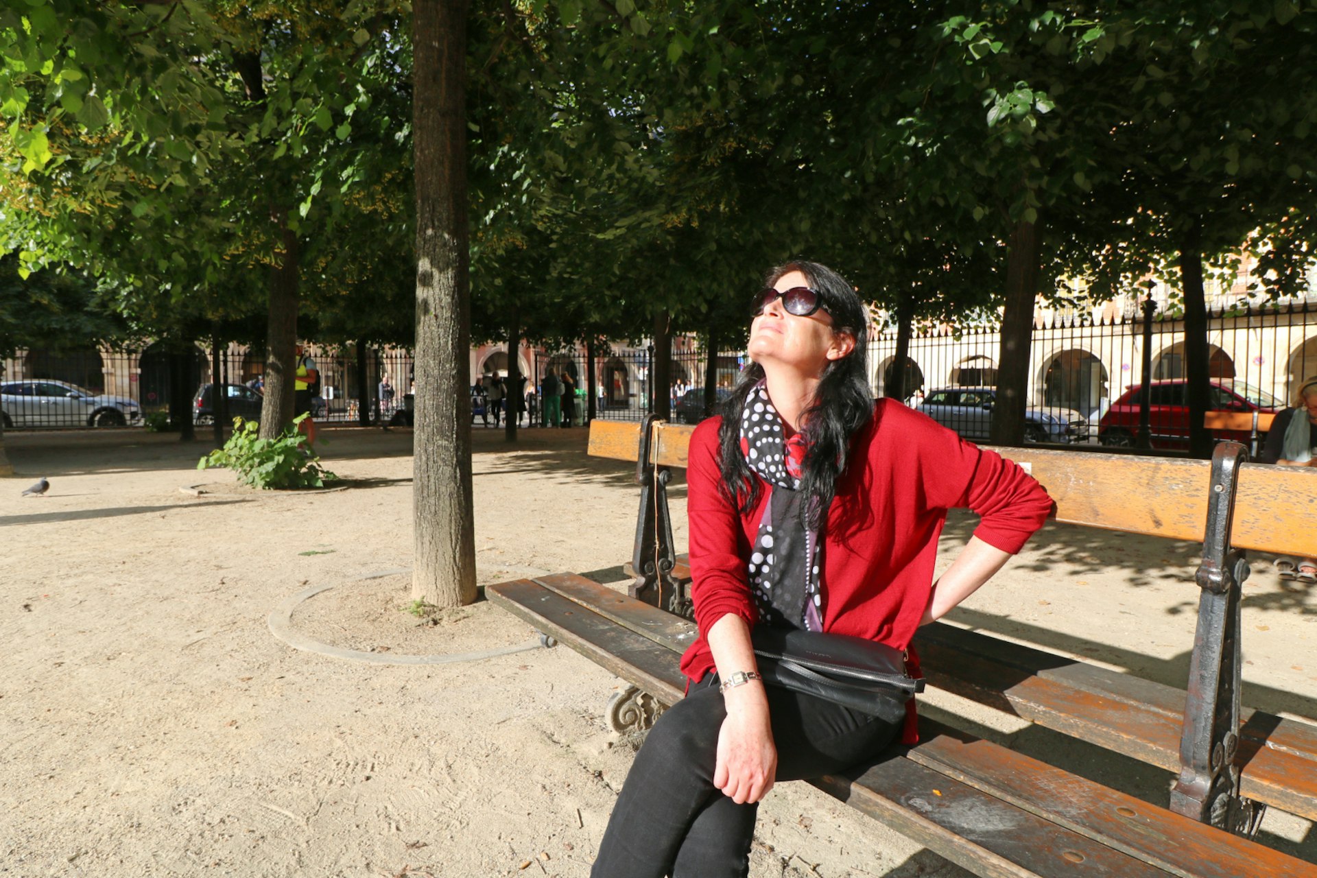 Lonely Planet writer Catherine Le Nevez soaks up the sun at place des Vosges in Paris, France; Catherine is sitting on a bench gazing up at the sun; she is wearing black trousers, a red cardigan and sunglasses; there are trees and iron railings behind her.