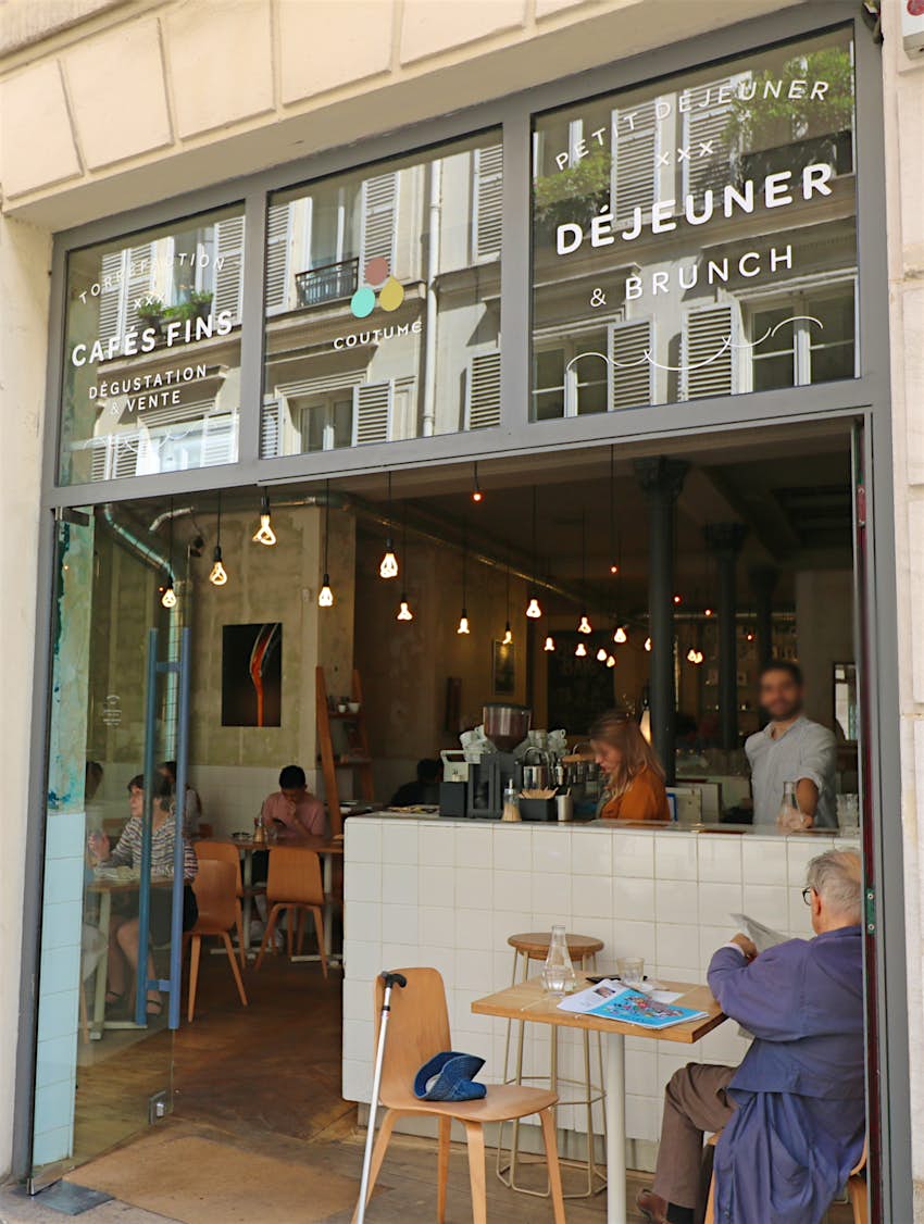 A man sits on a table outside of Coutume Café in Paris; the cafe has a modern glass front, with doors that have been fully opened to reveal a white tiled bar behind which servers are standing. 