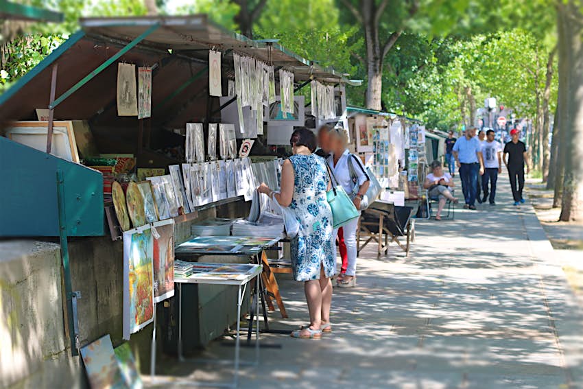 Customers browse the stalls of art dealers along the Seine in Paris, France; the stalls are arrayed under dark green shutters and there are trees in the background