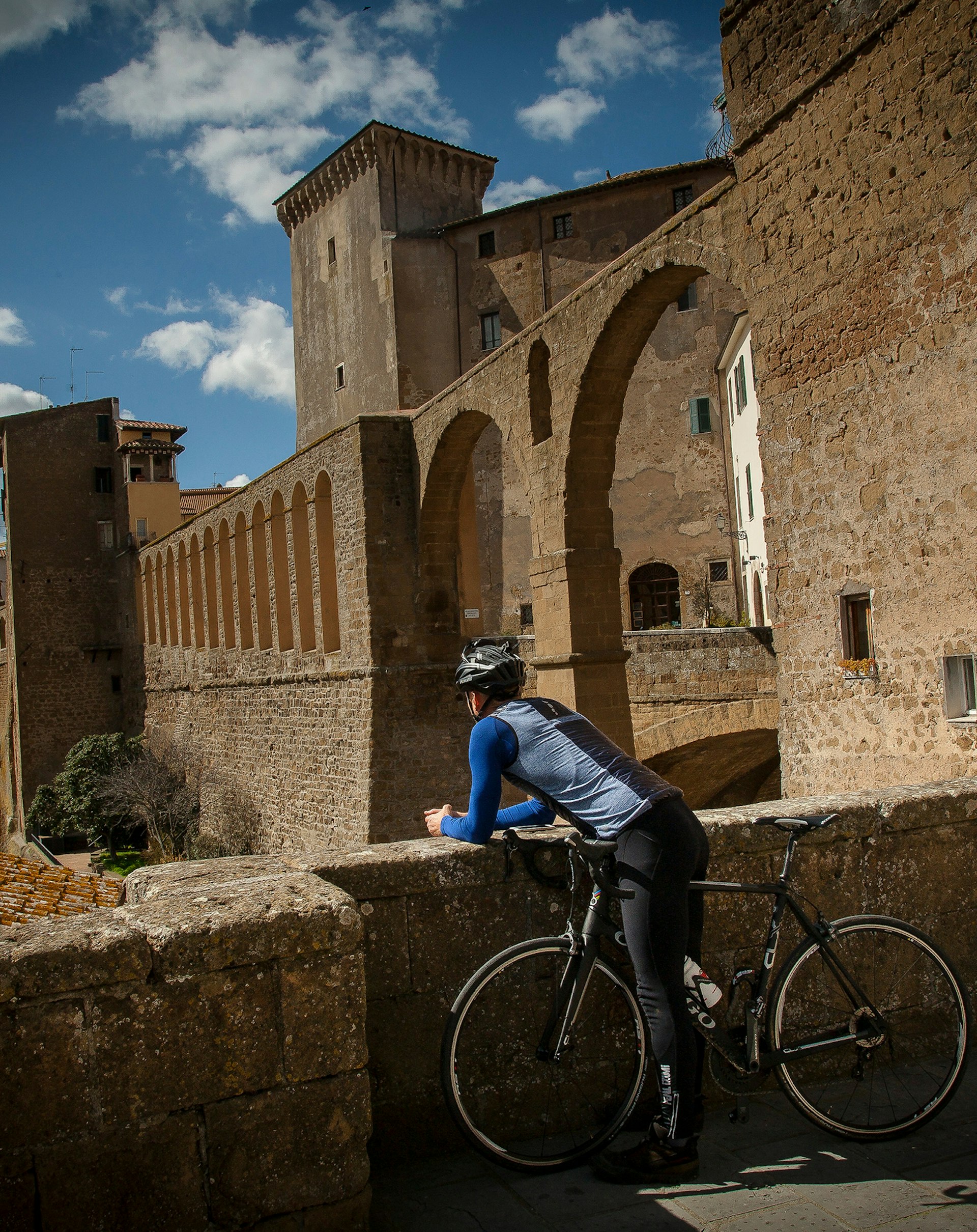 A cyclist leans over his handlebars onto a low wall and gazes out over a towering aqueduct with beautiful arches in the hilltop village of Pitigliano © Ciclica & Foto Mario Llorca.com