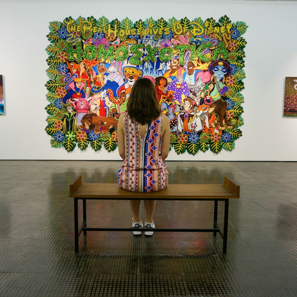 A woman sits in the middle of a simple bench with her back to the camera. In front of her are a number of artworks hanging on the wall, one featuring 'The Housewives of Disney', a colourful-cartoon painting © Monica Suma / Lonely Planet