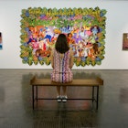 A woman sits in the middle of a simple bench with her back to the camera. In front of her are a number of artworks hanging on the wall, one featuring 'The Housewives of Disney', a colourful-cartoon painting © Monica Suma / Lonely Planet