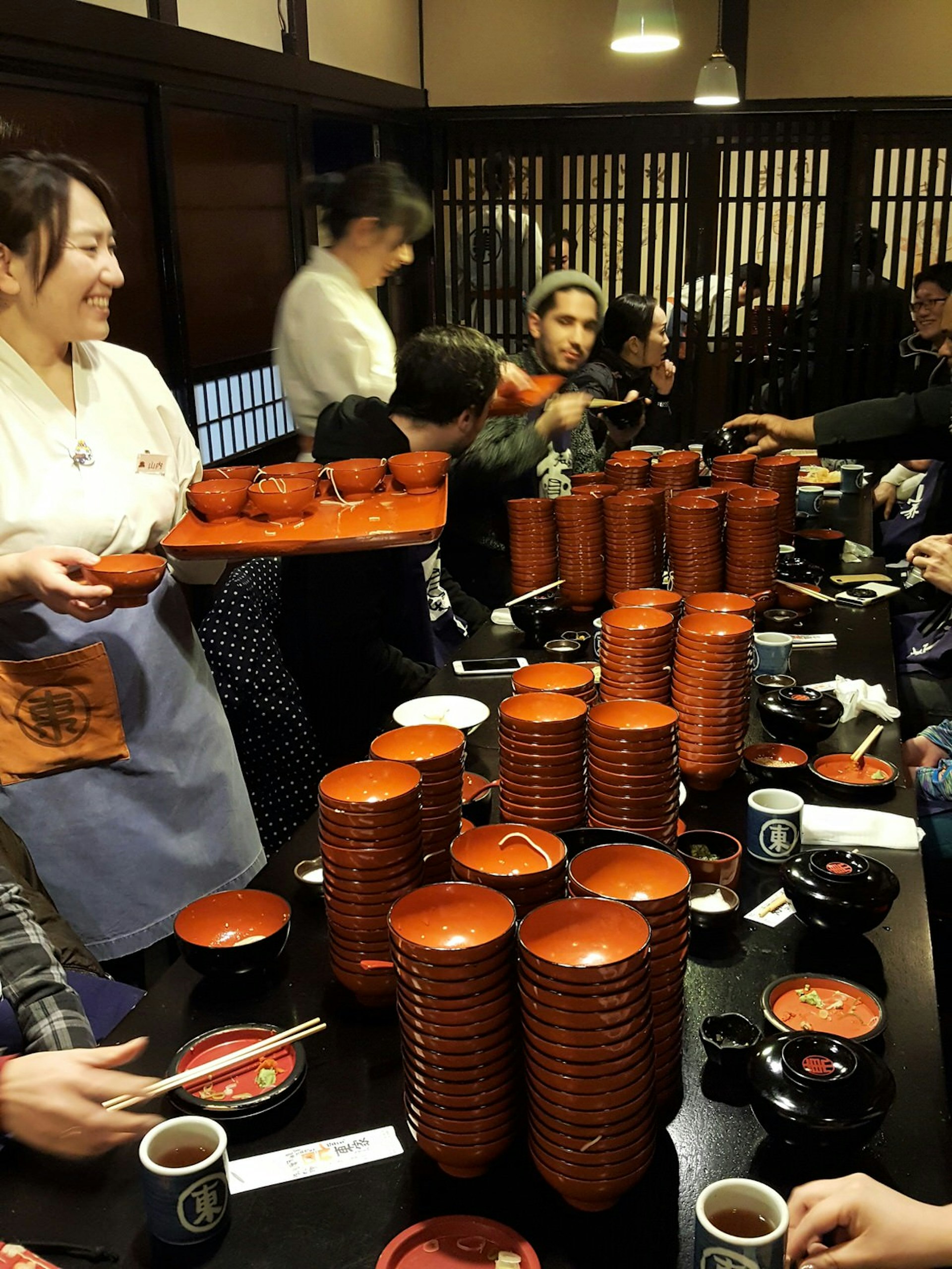 The busy interior of Azumaya, Morioka with long table full of stacks of small empty noodle bowls and customers, as staff stand beside with trays of more noodles