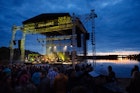 A large outdoor, open-air stage is lit at twilight as it sits beside a lake, while music fans listen © Festival de musique émergente