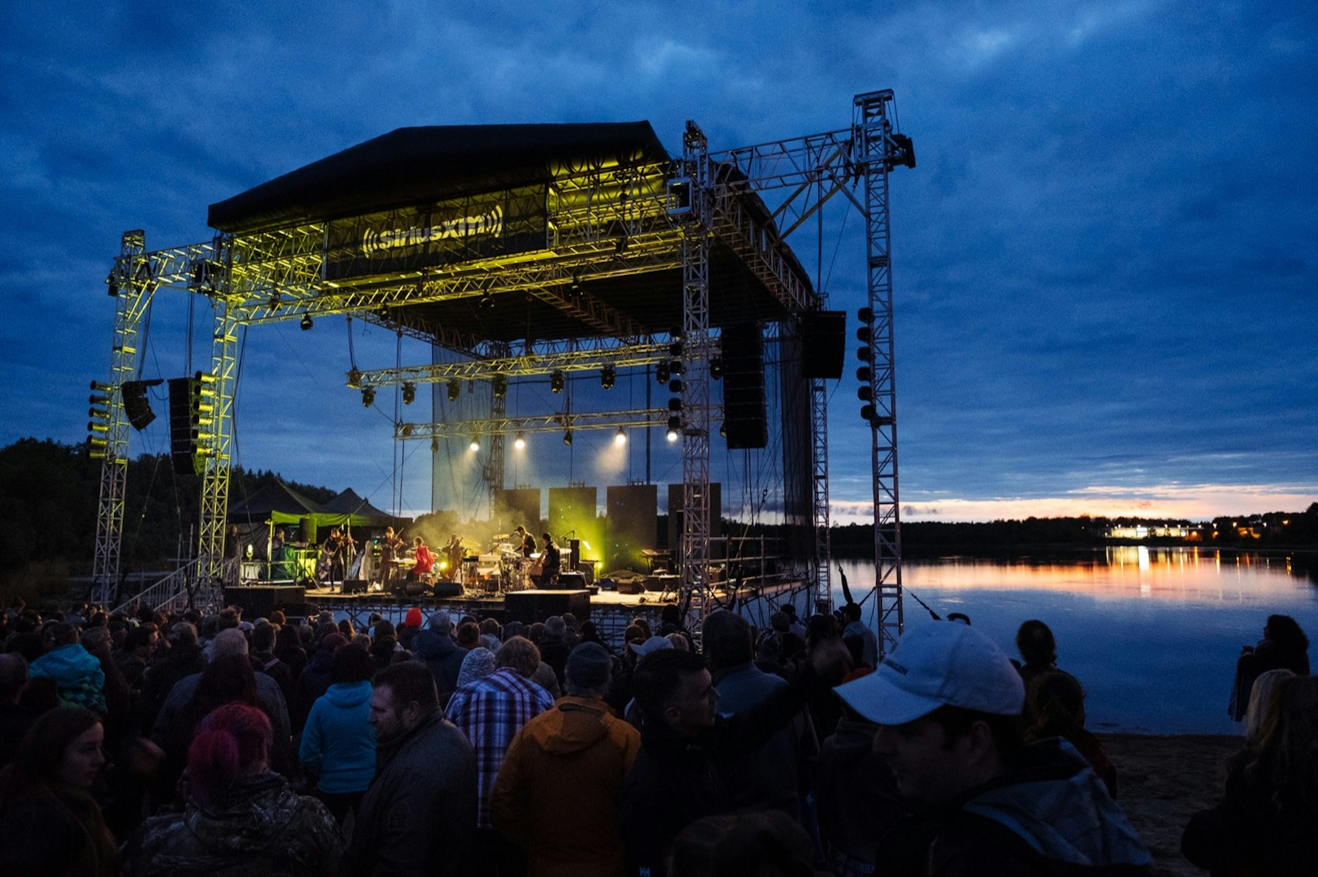 A large outdoor, open-air stage is lit at twilight as it sits beside a lake, while music fans listen © Festival de Musique Émergente