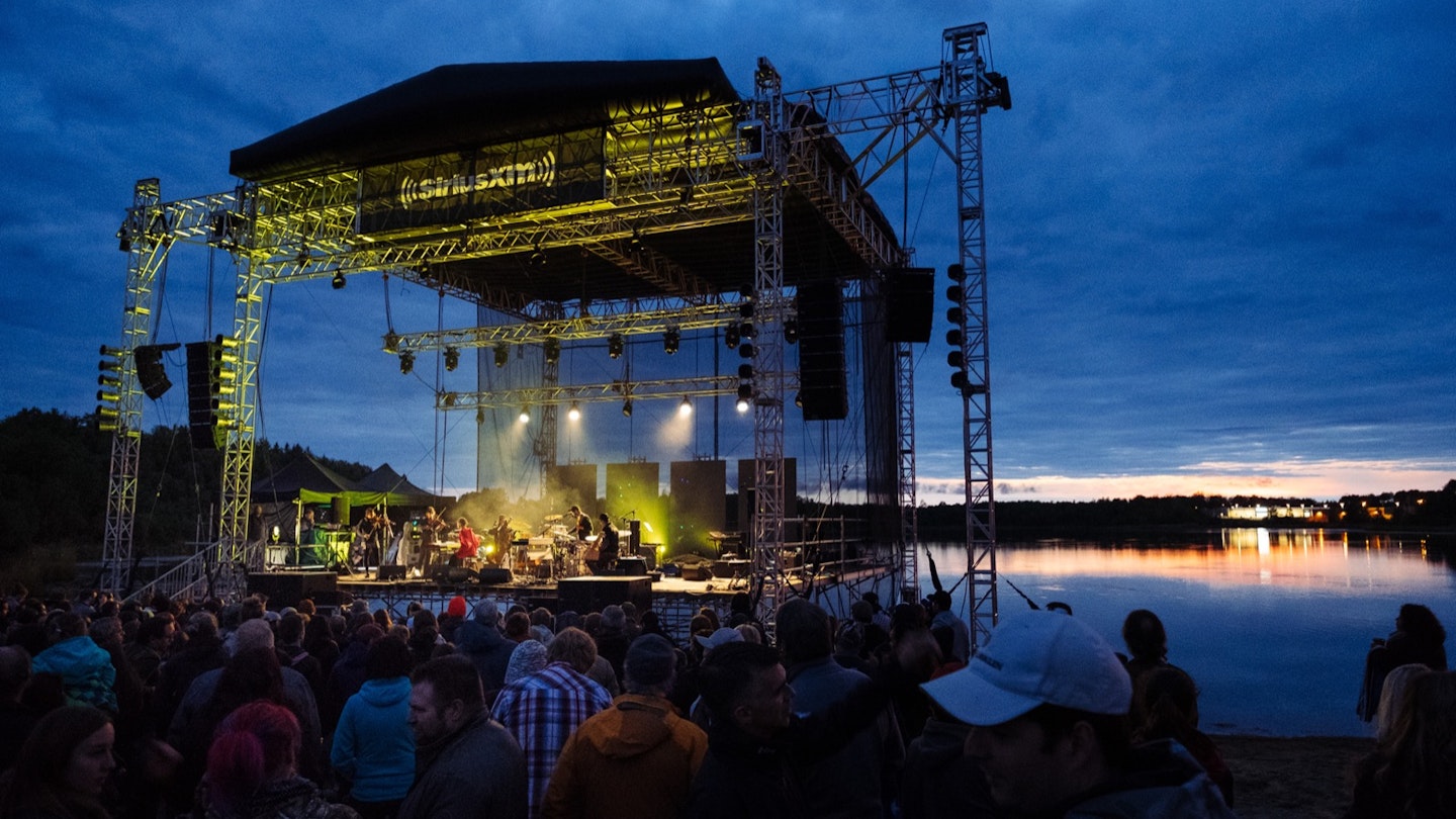 A large outdoor, open-air stage is lit at twilight as it sits beside a lake, while music fans listen © Festival de musique émergente