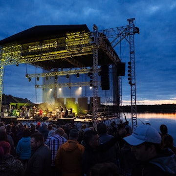 A large outdoor, open-air stage is lit at twilight as it sits beside a lake, while music fans listen © Festival de musique émergente
