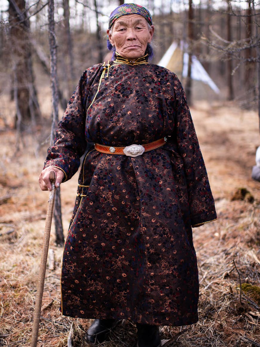 En äldre mongolisk kvinna i traditionell klädsel står i en vintrig skog.  Möte med en medlem av en lokal renstam i Mongoliet © Chris Sheldrick