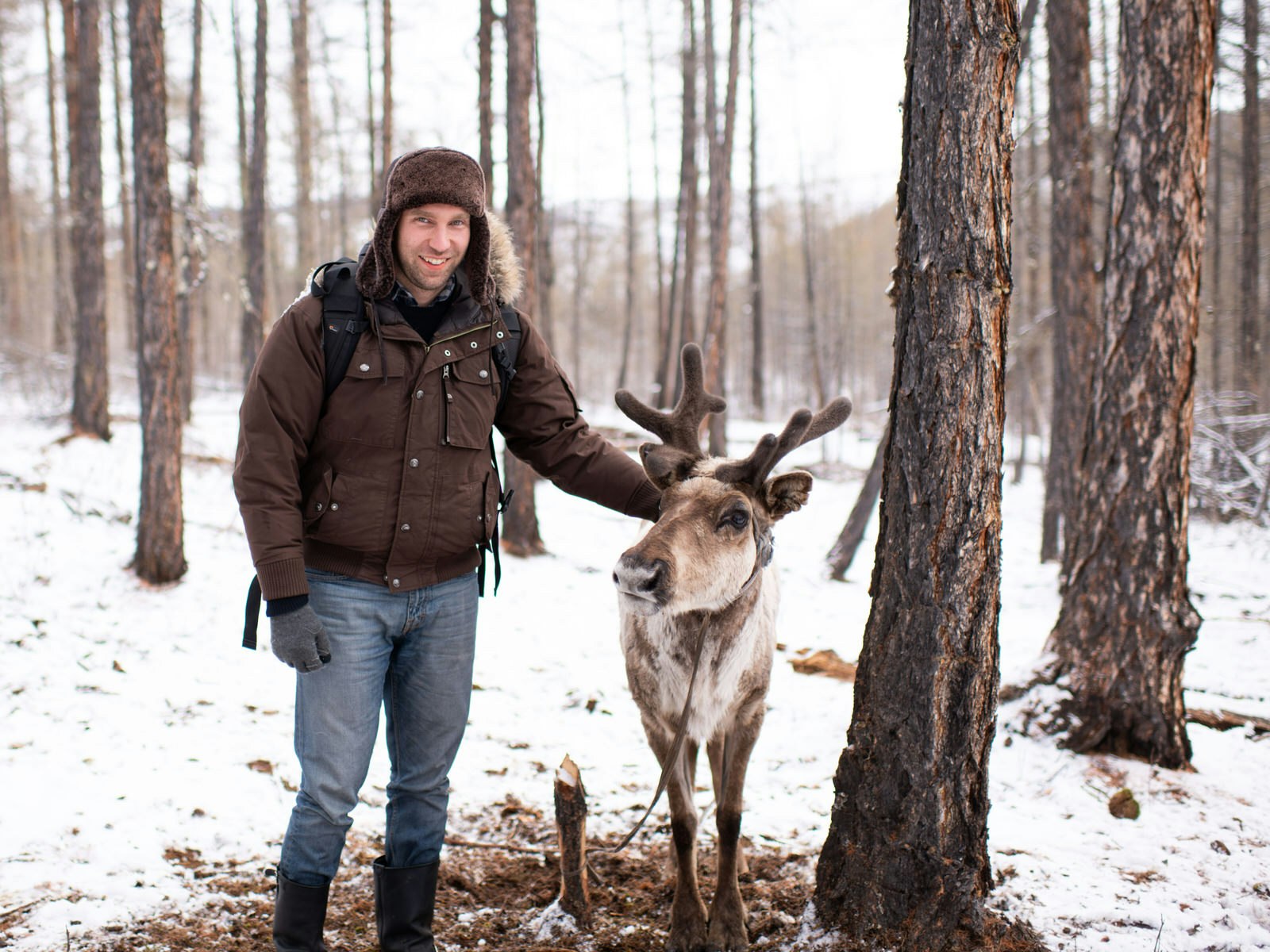 Chris Sheldrick wearing a fur hat and winter coat pets a reindeer in a snowy forest. Despite a packing faux pas, Chris was able to make friends in northern Mongolia © Chris Sheldrick