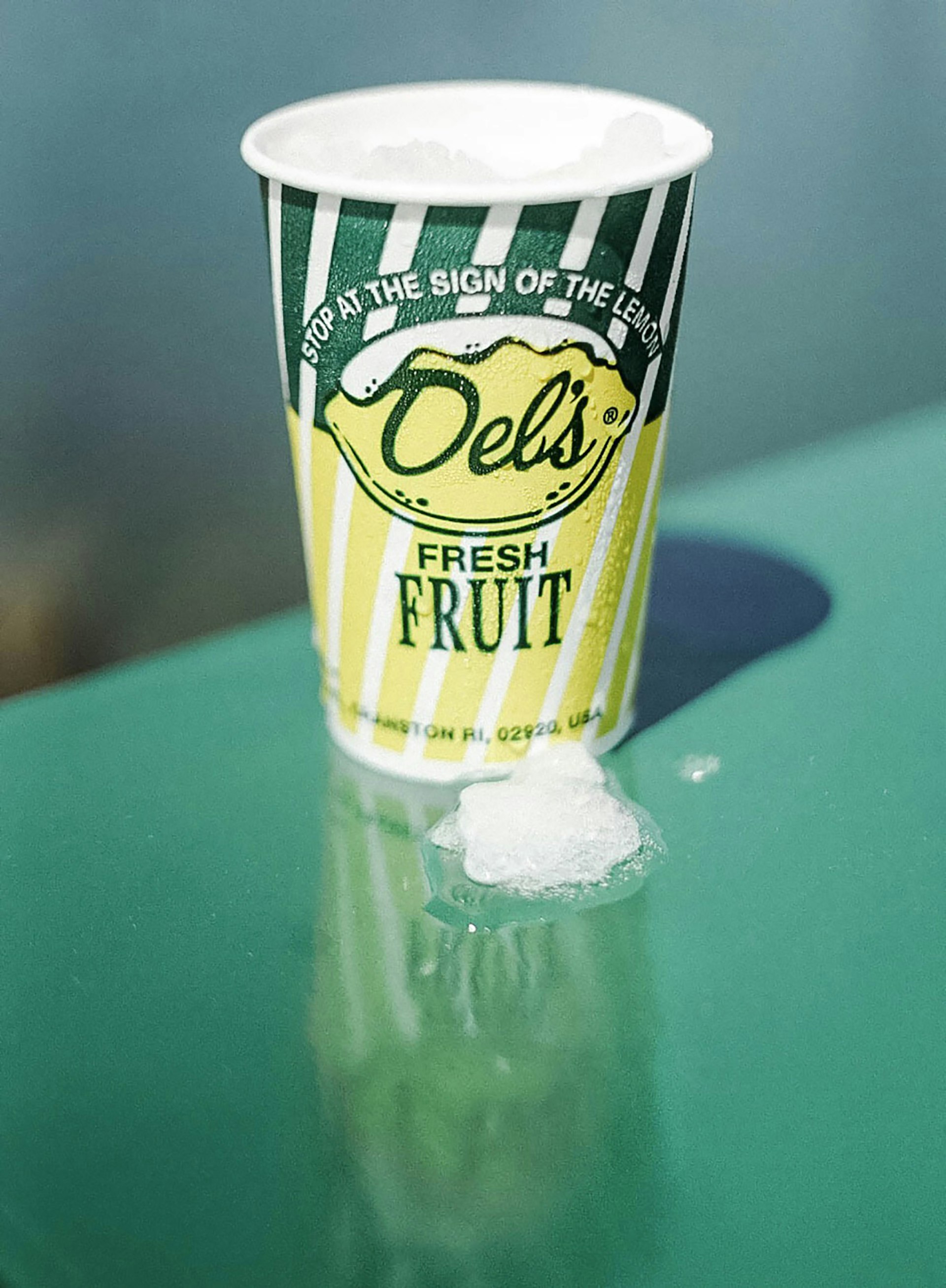 Close-up of a paper cup with vintage yellow and green logo for Del's Fresh Fruit lemon ice in Rhode Island, on a green table © Anna Saxon / Lonely Planet