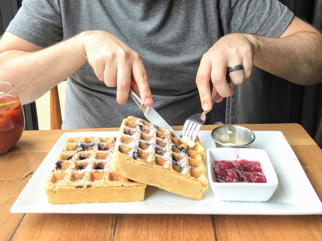 Close-up of man in gray t-shirt cutting into two stacked waffles on a white plate, with a side of jam © Cate Huguelet / Lonely Planet