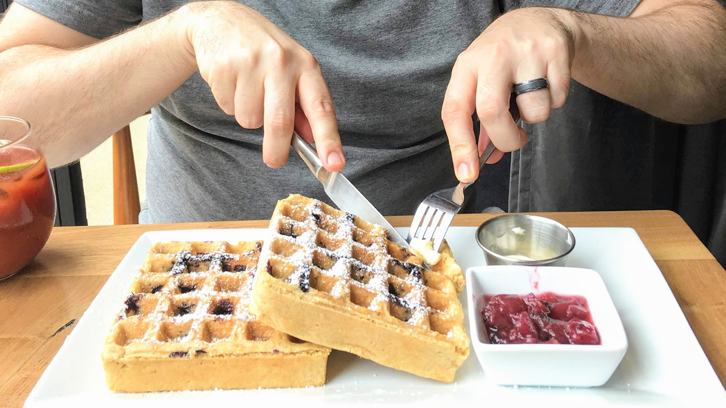Close-up of man in gray t-shirt cutting into two stacked waffles on a white plate, with a side of jam © Cate Huguelet / Lonely Planet