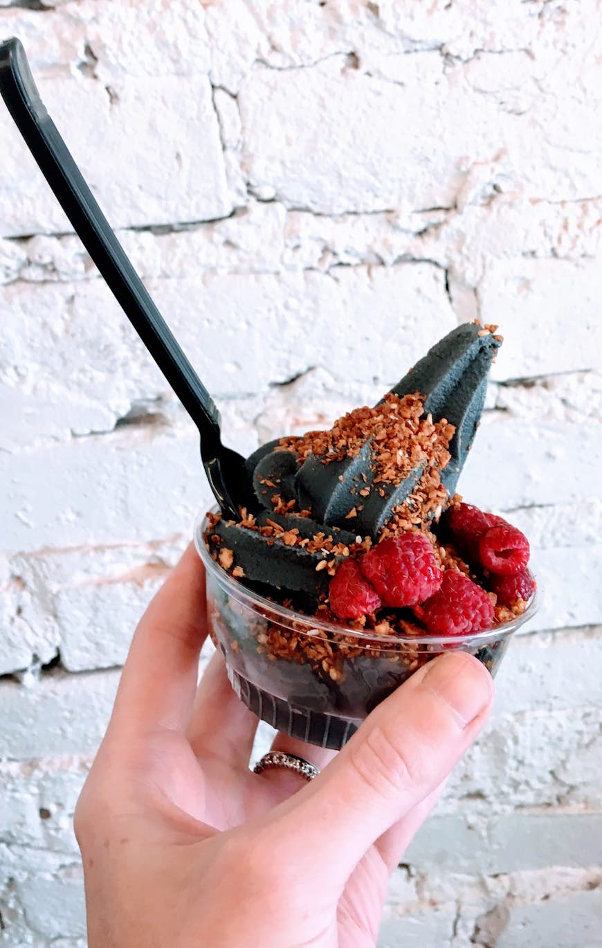 Close-up of a hand holding small plastic cup of chocolate FroYo topped with berries, with a white brick wall as backdrop; healthy chicago