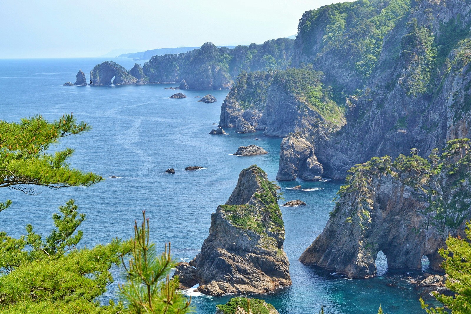 View of the rocky coastline of the Kitayamazaki Cliffs in Iwate on a clear and sunny day. The sheer, rocky cliffs are topped with bright green grass and surrounded by restless sea.