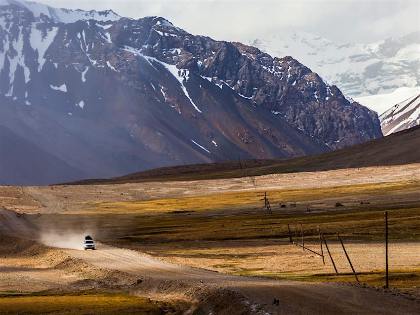 Features - Chris's bucket list travel experience: a road trip down the Pamir Hwy through Tajikistan ©NOWAK LUKASZ/Shutterstock A 4WD drives down a dirt road through a valley surrounded by mountains. Chris's bucket list travel experience: a road trip down the Pamir Hwy through Tajikistan ©NOWAK LUKASZ/Shutterstock