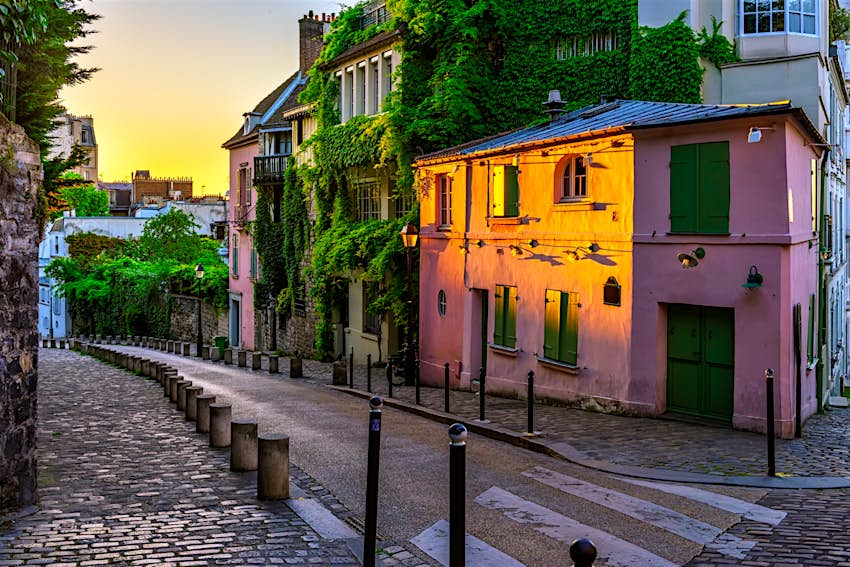 The sun sets over the Montmartre quarter in Paris, France; we are looking down a deserted street, where a pink house is bathed in sunlight, and the adjoining property is covered in ivy