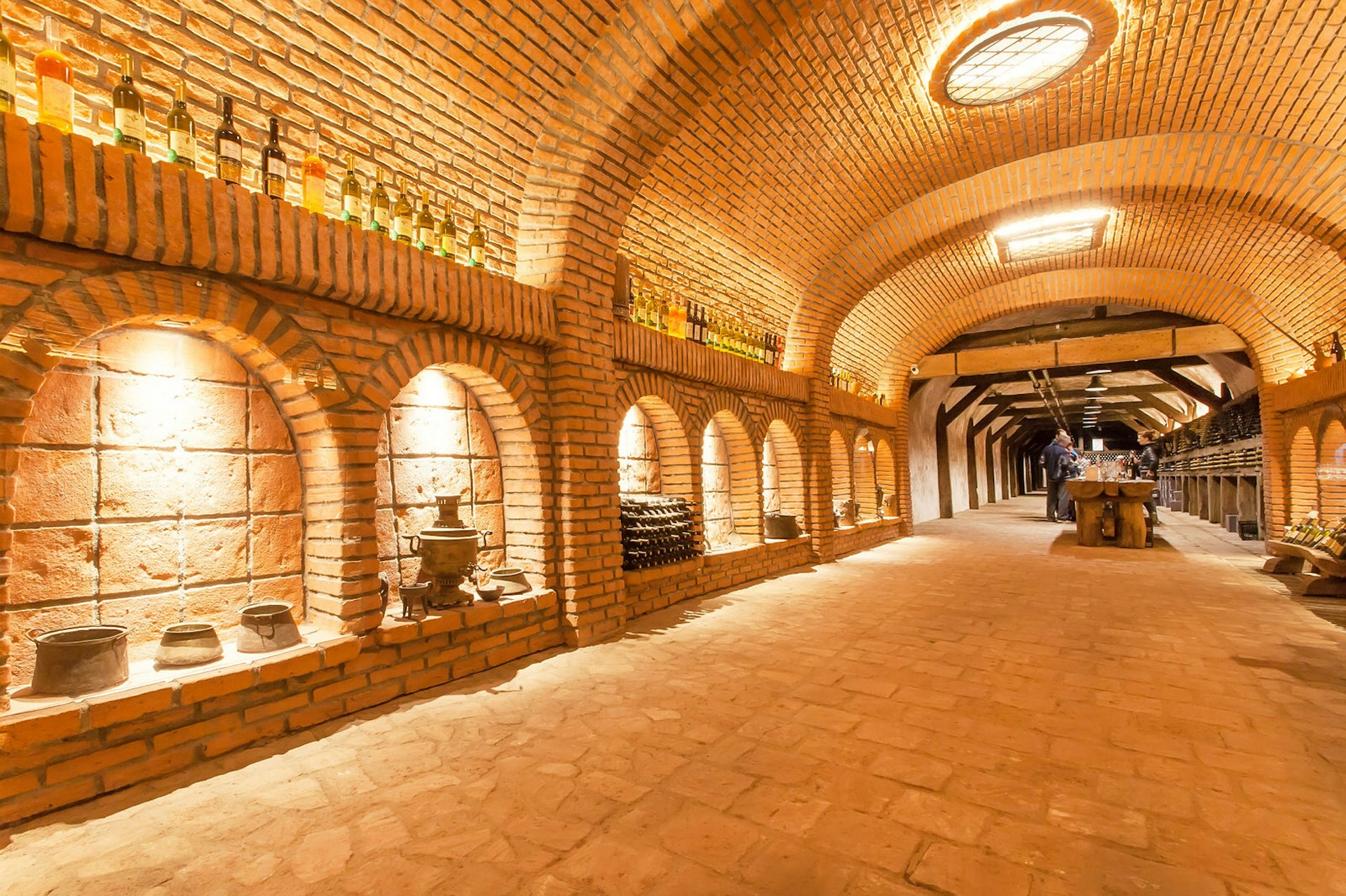 A well-lit tunnel with vintage wine-making equipment and wine bottles on display with a wine-tasting session taking place in the background at Winery Khareba, Georgia © Radiokafka / Shutterstock