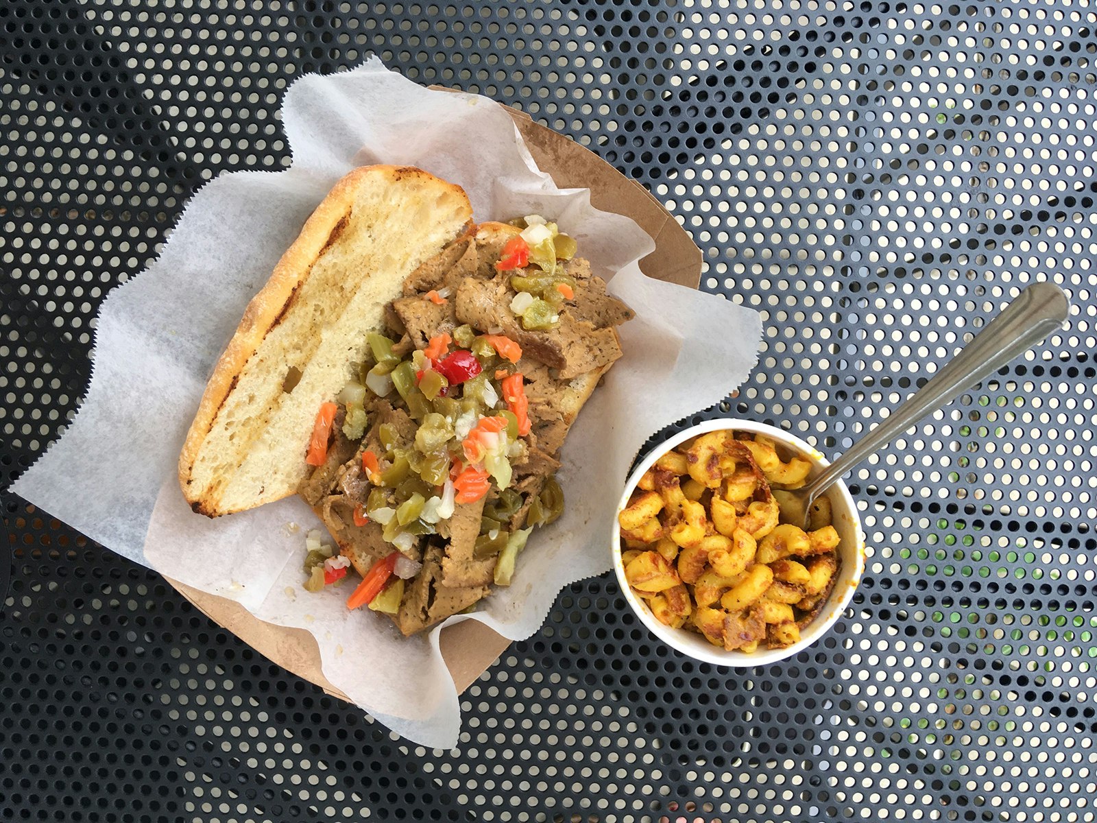 Vegan version of an Italian beef sandwich, and cup of vegan mac-and-cheese, shot from above and arrayed on a black metal table; healthy chicago