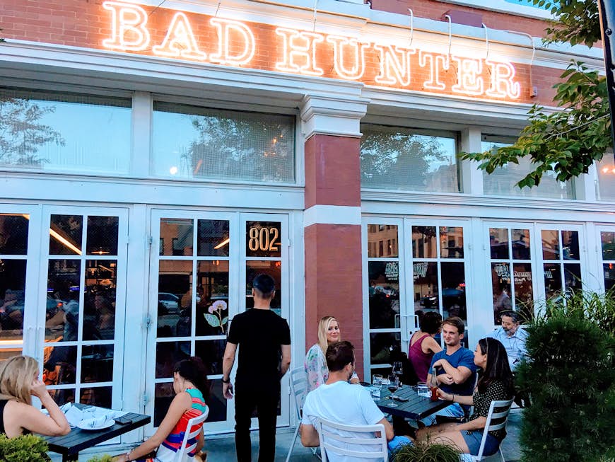 Exterior shot of Chicago restaurant Bad Hunter, with white French doors, neon signage and a small patio filled with customers; healthy chicago