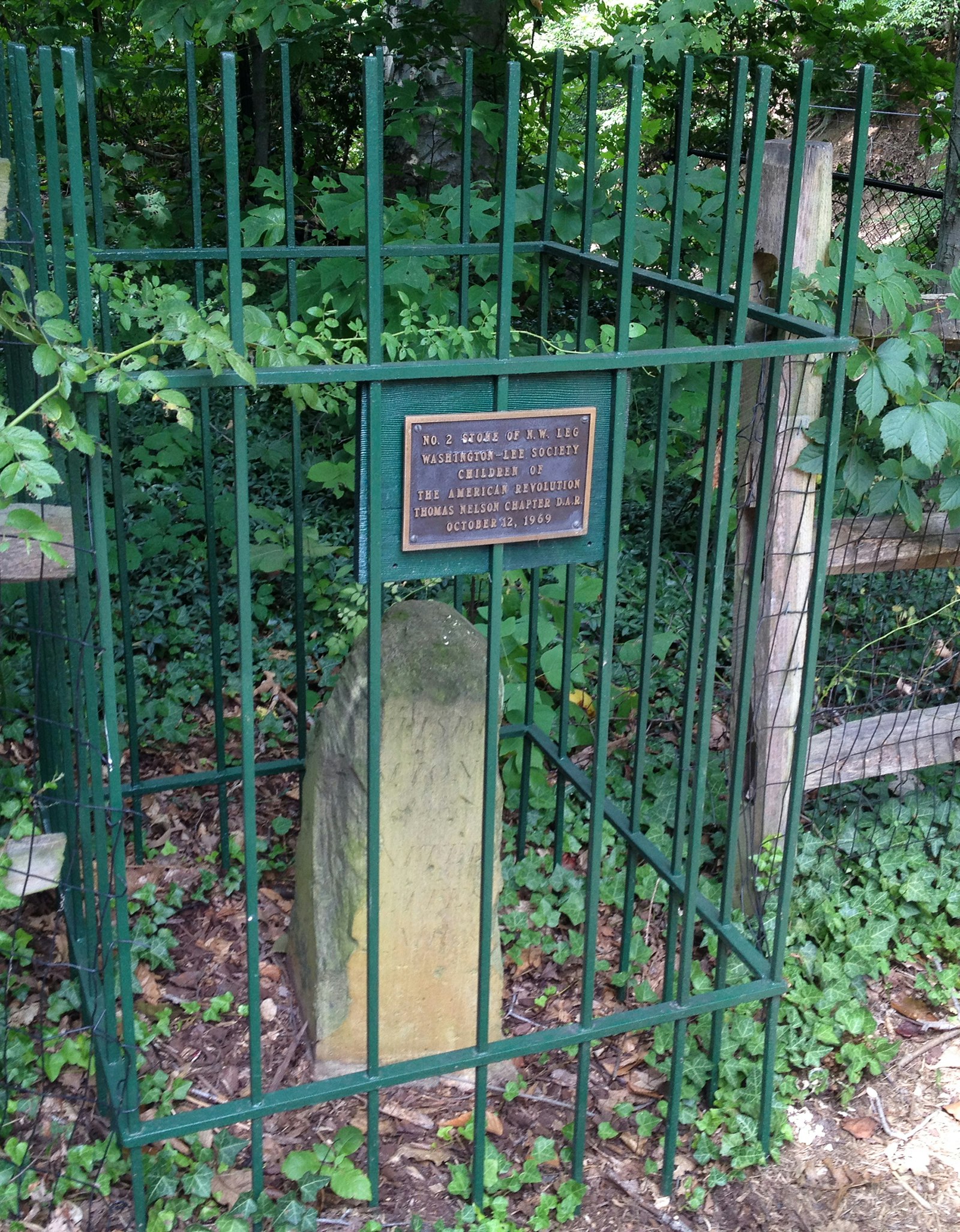 A three-foot stone with a rounded top sits behind a green fence marked by a plaque that explains the stone is one of 40 that originally established the city's boundaries. Green vines form the backdrop. © Barbara Noe Kennedy / Lonely Planet
