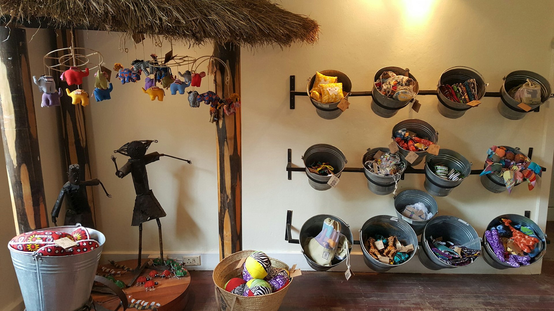 An image of the store interior, with a neat rows of large tin buckets hung on the wall, each holding colourful children's toys made of brightly-coloured fabric. In the corner, beneath a thatched gazebo of sorts, hang lovely mobiles of little fabric elephants. Below them are little tin statues of people made from old cans. Pefect Nairobi shopping for kids at home © Clementine Logan / Lonely Planet