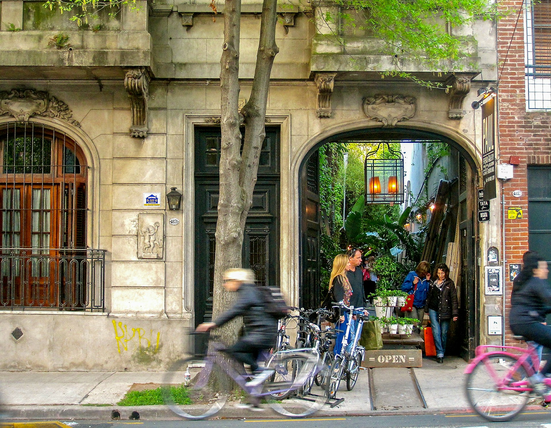 Street scene of people coming out of vine-covered alley with two cyclists zipping by on the street © Bridget Gleeson / Lonely Planet