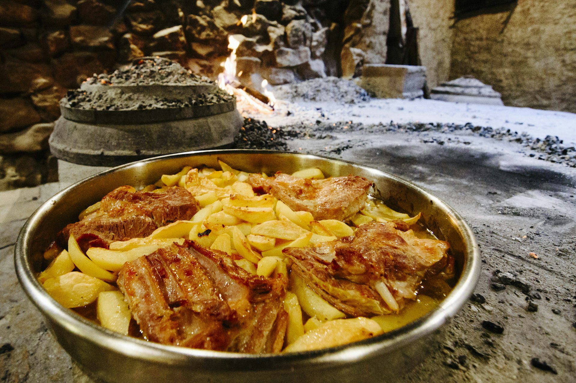 A metal dish containing meat and potatoes, with the ashes of a fireplace and the peka lid in the background © Wine & Food Hedonism