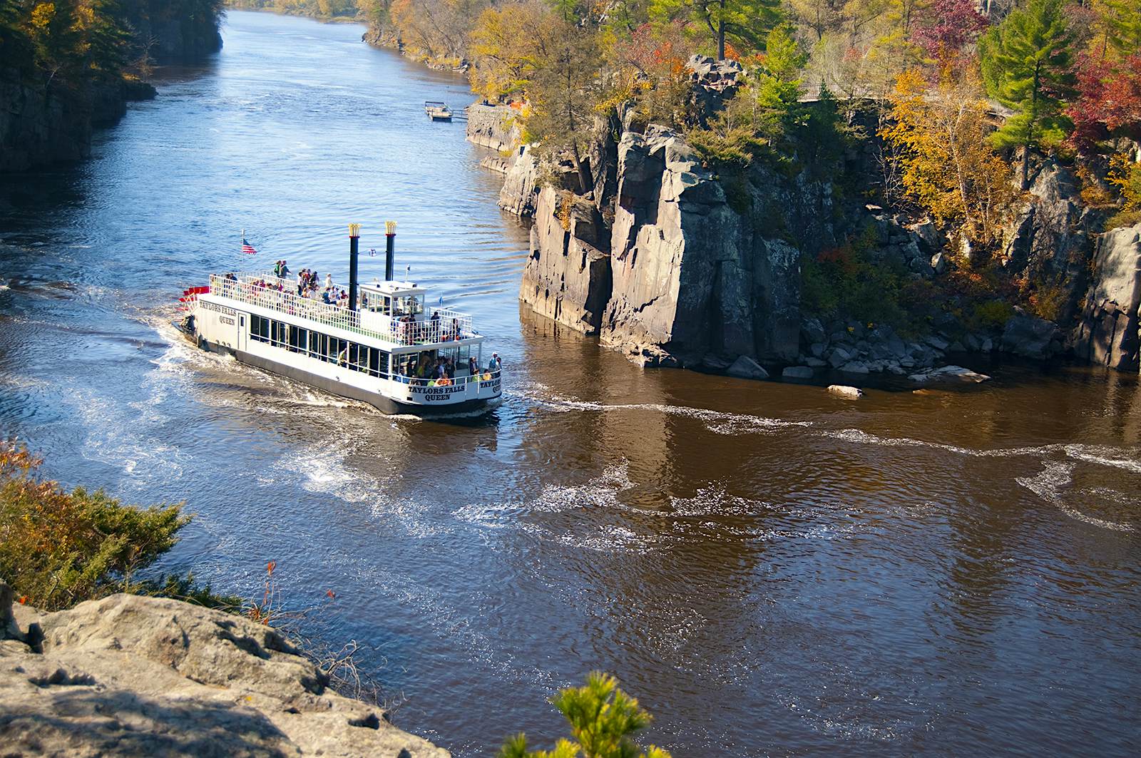 A paddle boat on the St. Croix River. Trees on the rocky riverbanks have leaves of red, orange, gold and green