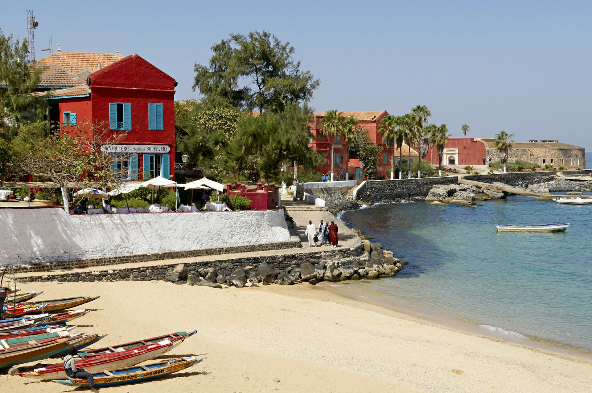 Looking out from the beach on Gorée to the sea, the left side of the image has a couple of traditional piroques on the sand, which are backed by bright red historical buildings (two storeys) and trees © Tuul & Bruno Morandi / Getty Images