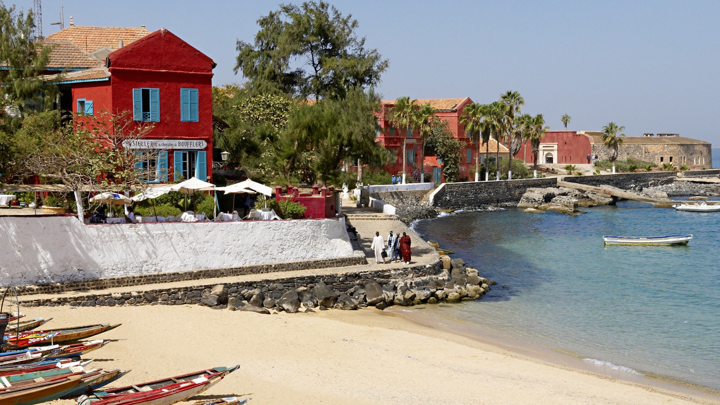Looking out from the beach to the sea, the left side of the image has a couple of traditional piroques on the sand, which are backed by bright red historical buildings (two storeys) and trees © Tuul & Bruno Morandi / Getty Images
