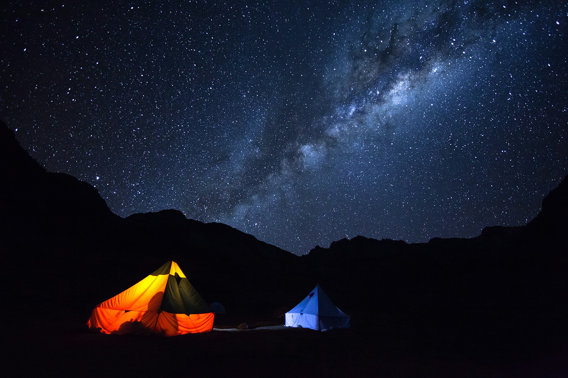 A nighttime shot of two lit-up tents in the foreground with a starry sky in the background (Milky Way visible) © Kesterhu / Getty Images