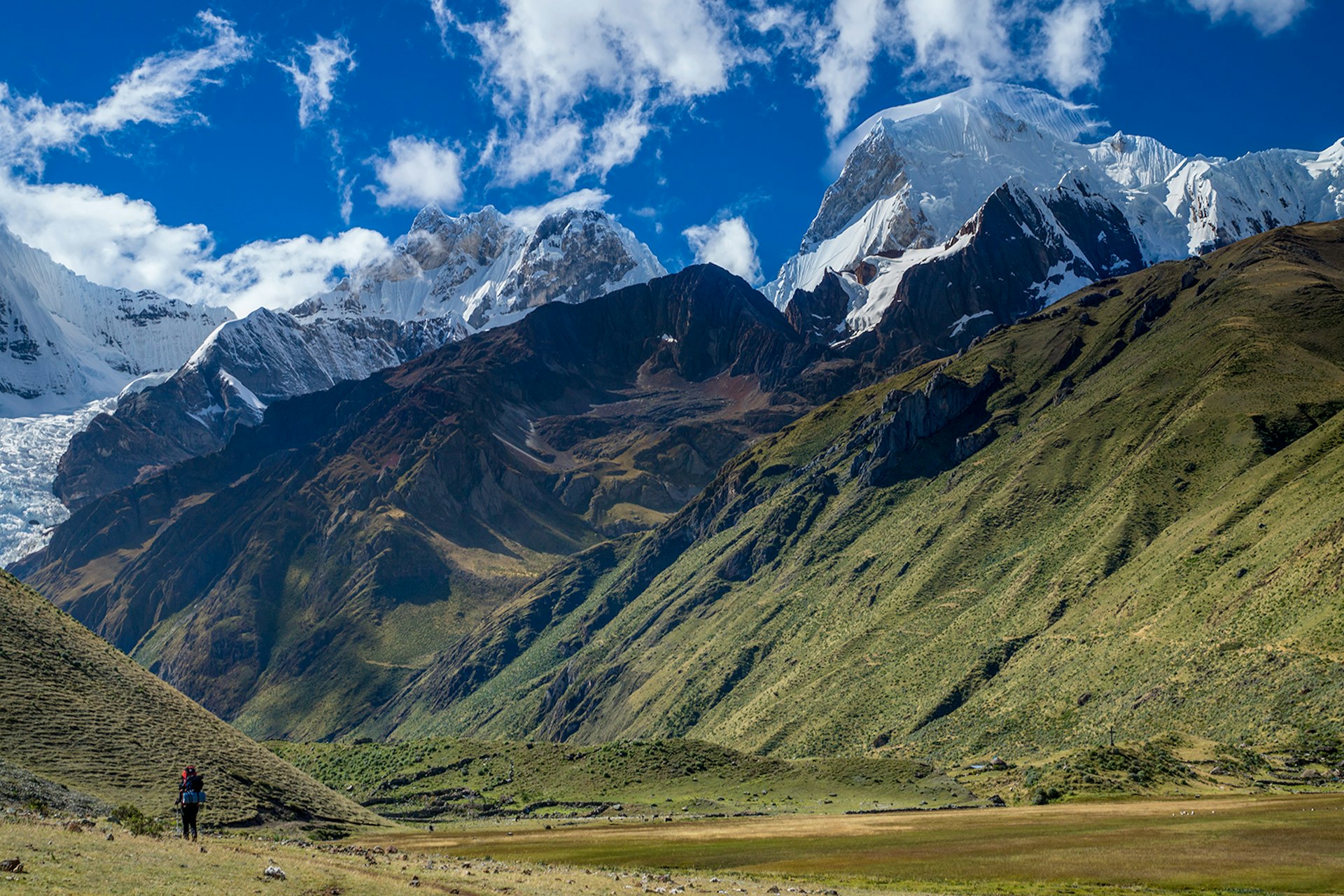 A trekker walks with green hills and rocky peaks in front of them © Diamirstudio / Getty Images